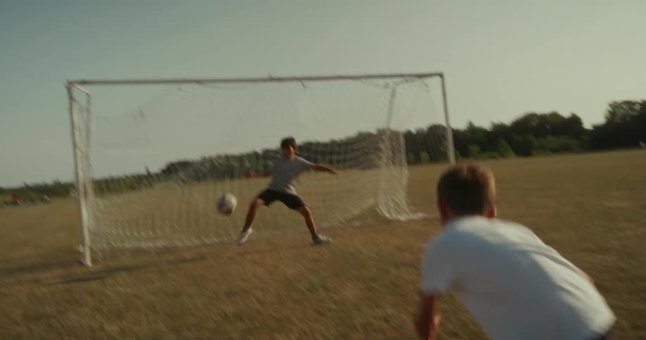 A lowangle shot of a young boy running on a grassy field dribbling a soccer ball he kicks the ball towards a soccer goal where another person likely a goalkeeper is waiting