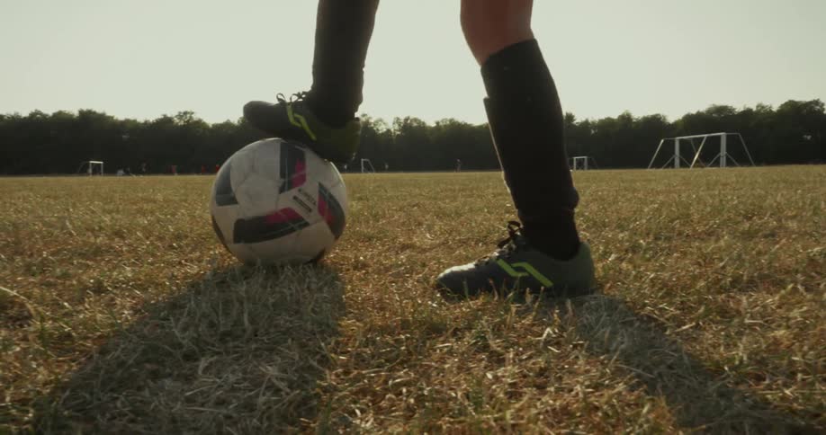 A soccer ball being kicked with the focus on the ball and the movement of a persons legs and feet the setting appears to be a dry grassy field with a soccer goal visible in the background