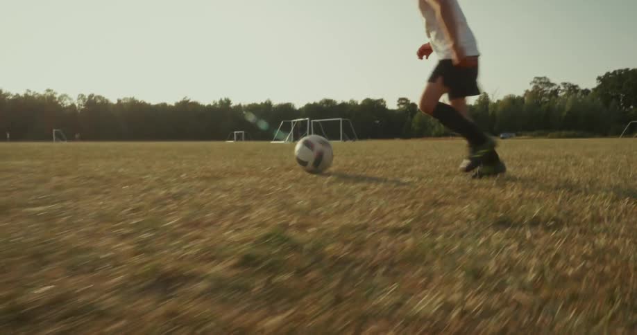 A child kicks a soccer ball toward the goal while another child stands ready as goalkeeper on a sunlit dry field