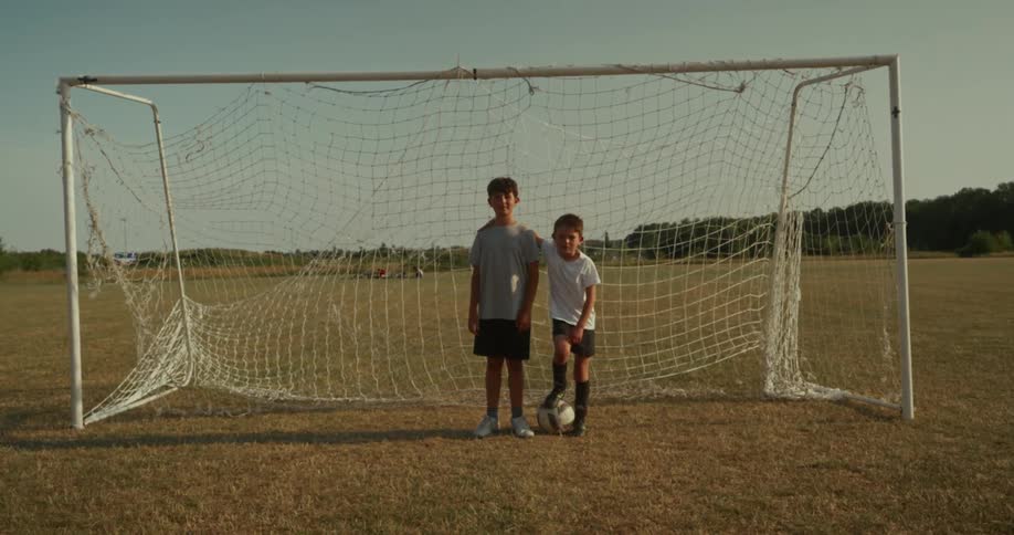 Two friends hugging in a soccer goal with one of them holding a ball with his feet