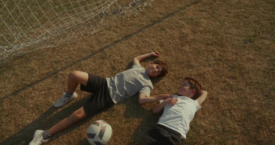 Two young boys are lying on a football field at midday talking to each other with a soccer ball between them