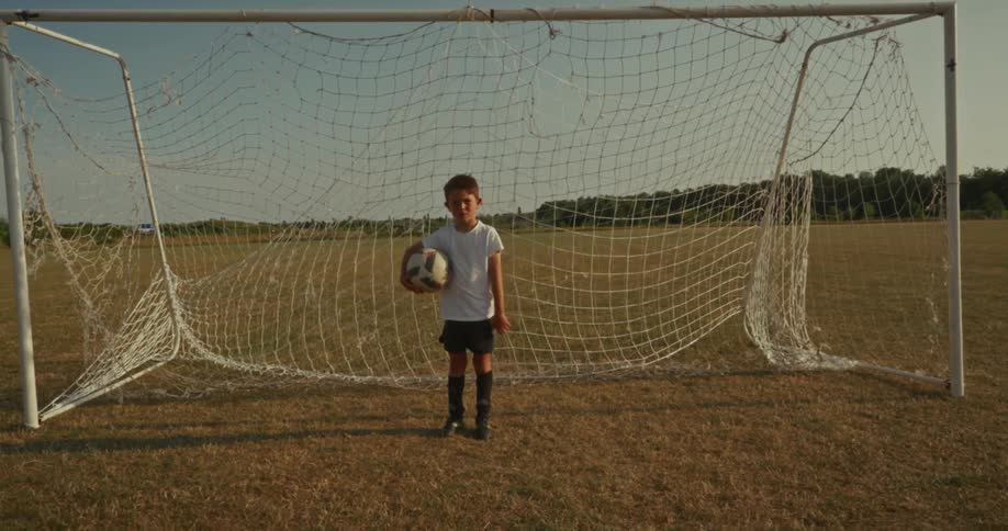 A young child stands in front of a soccer goal on a grassy field holding a soccer ball