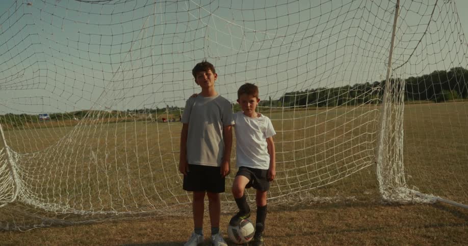 Two young boys standing in front of a soccer goal on a dry grassy field under a bright sky both boys are dressed in athletic wear and a soccer ball is positioned between them resting near the foot of the shorter boy