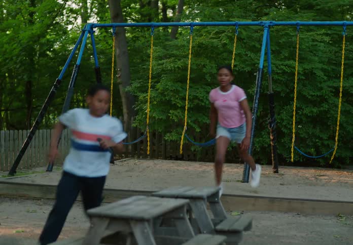 A teen girl and a boy both on the play area with the girl sitting on a swing gaming as the boy took the handheld game from her and run as the girl chase after him
