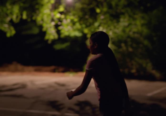 A teenage girl walks alone in the middle of a quiet residential street at night with streetlights casting a soft glow and houses lining either side