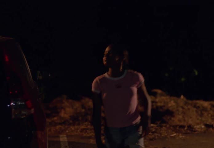 A girl in a pink shirt and white shoes walks towards a red car at night she kneels down to examine something on the ground near the car then stands up and looks around thoughtfully