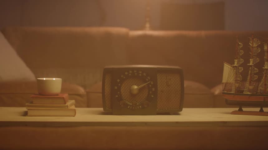 A hand places a cup on a stack of books beside a lit candle and model ship near a vintage radio on a wooden coffee table in front of a brown leather couch