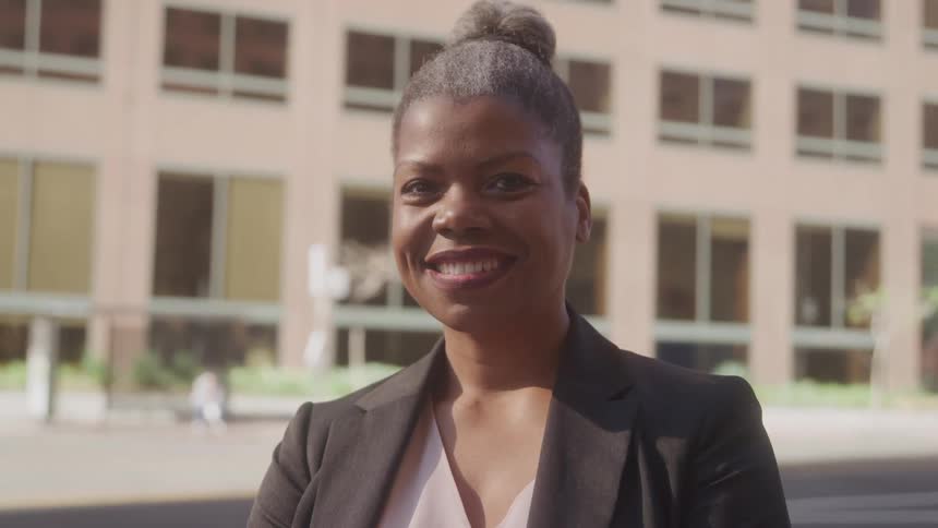 A black woman wearing a black suit with a ponytail and a smile on her face is standing beside an office building at midday