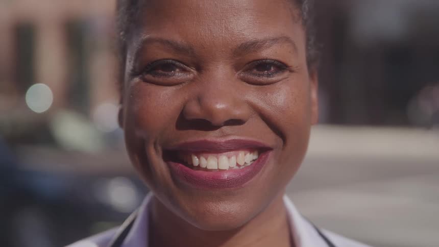 A woman in a black blazer and white shirt stands in front of busy street smiling directly at the camera
