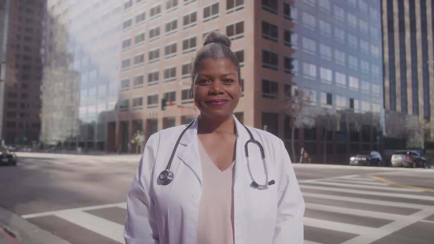 A doctor with a stethoscope around her neck stands in a city street looking at the camera with tall buildings behind her