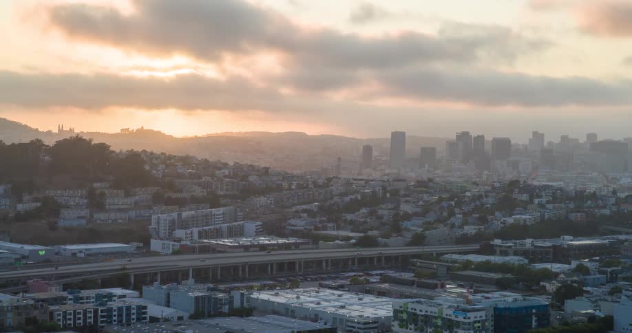 An aerial view showcases a sprawling urban landscape under a warm hazy sky at sunset with sunlight breaking through scattered clouds a multilane highway cuts through the middle ground surrounded by a mix of residential and commercial buildings