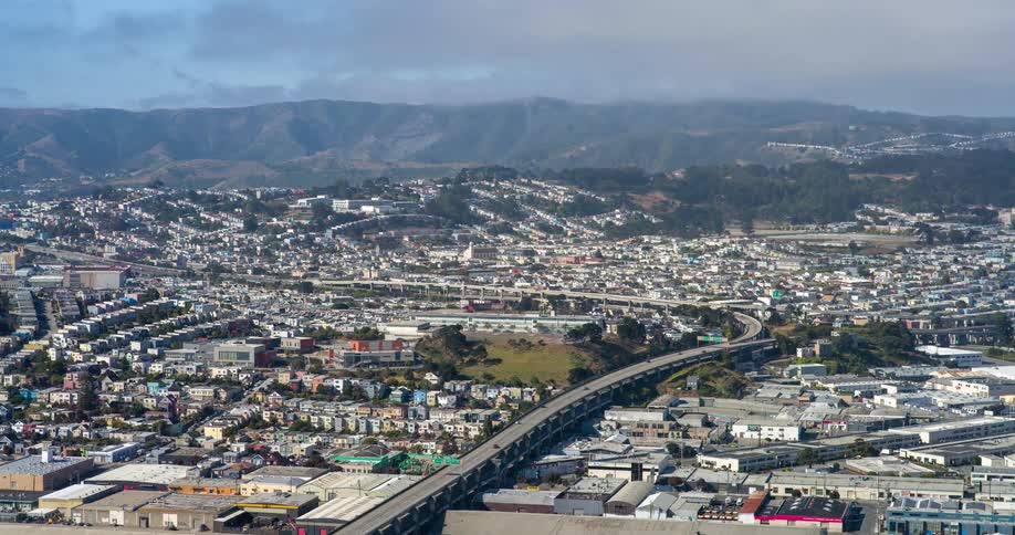 An aerial drone shot captures a densely populated urban area with a highway snaking through a valley between numerous buildings the surrounding landscape features rolling hills suggesting the sprawl of a major city