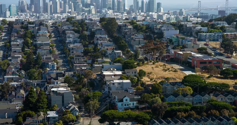 San franciscos skyline rises in the distance beyond a dense grid of residential rooftops with the bay bridge stretching across the right side of the frame