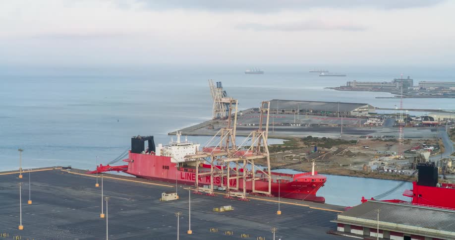 A large red cargo ship is docked at a busy industrial port surrounded by cranes and warehouses with the ocean stretching into the distance