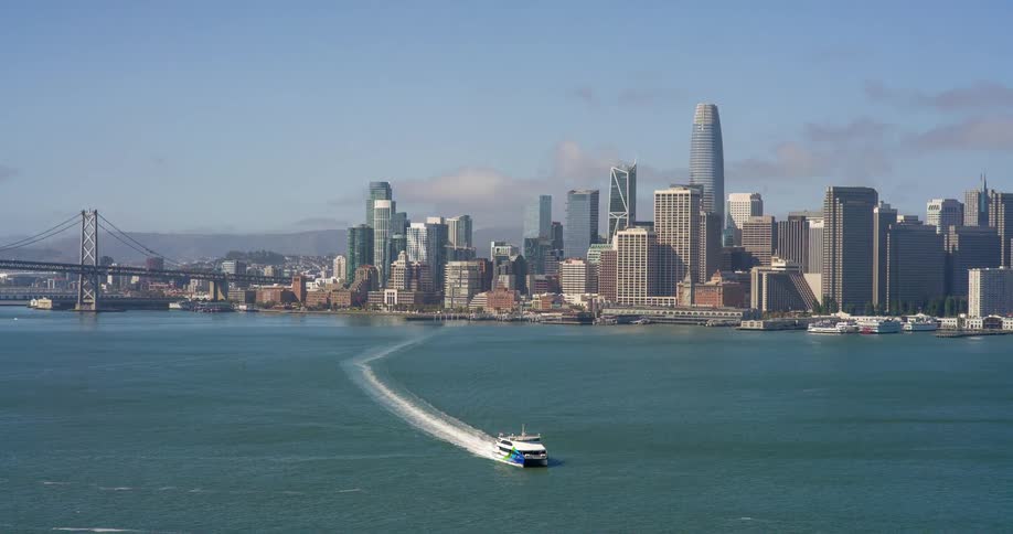 A ferry sails across the bay away from san franciscos skyline with the bay bridge stretching into the frame from the left