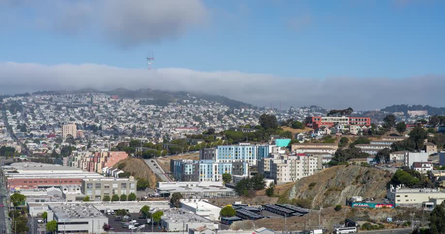 A dense hillside cityscape features residential buildings and towers clustered beneath a cloudshrouded radio antenna