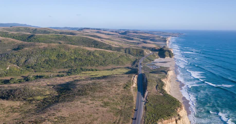 An breathtaking aerial view captures a long winding coastal highway stretching along a dramatic cliffside with the vast expanse of the pacific ocean and its crashing waves on the right rolling green hills flank the left side of the road
