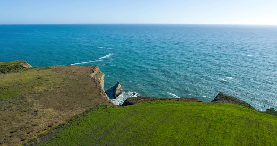 A grassy cliff juts above a calm blue ocean under a clear sky with rocky promontories scattered along the edge