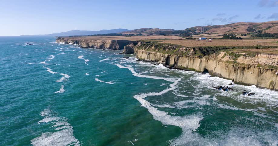 A rugged coastline with steep light brown cliffs meets a turquoise ocean with white waves crashing onto the shore