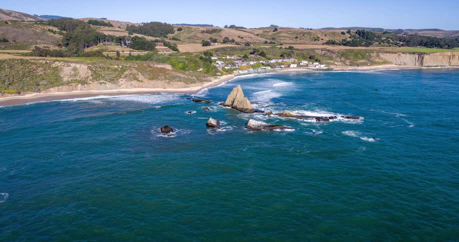 Aerial shot of rocks in the ocean at the edge of a beach with a small town in the background