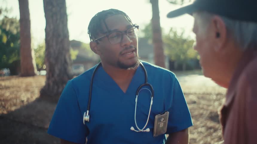 Two men one of whom is a doctor wearing a blue coat glasses and a stethoscope around his neck are talking in a park at midday the other man is elderly