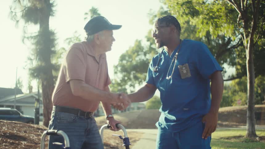 An elderly man using a walker shakes hands with a healthcare worker in blue scrubs outside near trees and a house