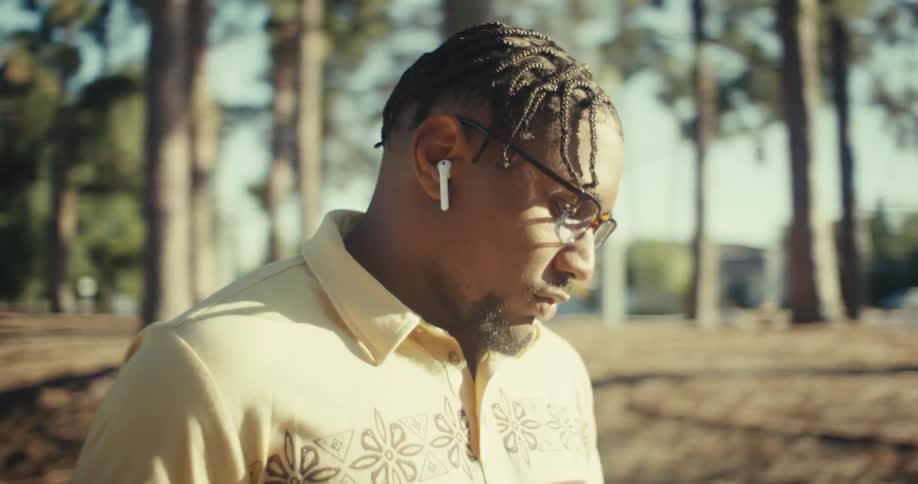 A man with braided hair and glasses wearing a yellow patterned polo shirt and white wireless earbuds he walks slowly through a park with tall trees suggesting he is enjoying a leisurely stroll while listening to something