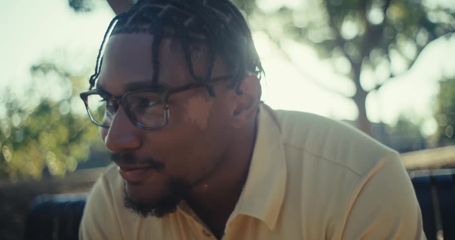 A young man with glasses and braids smiles and looks away seated on a bench in a sun dappled park