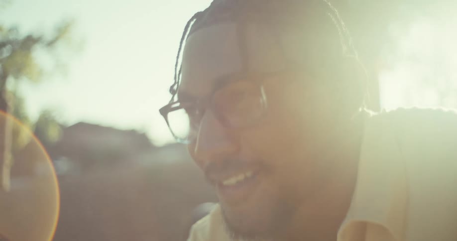 A happy black man sits focused on his phone while seated on a park bench beneath trees and soft daylight