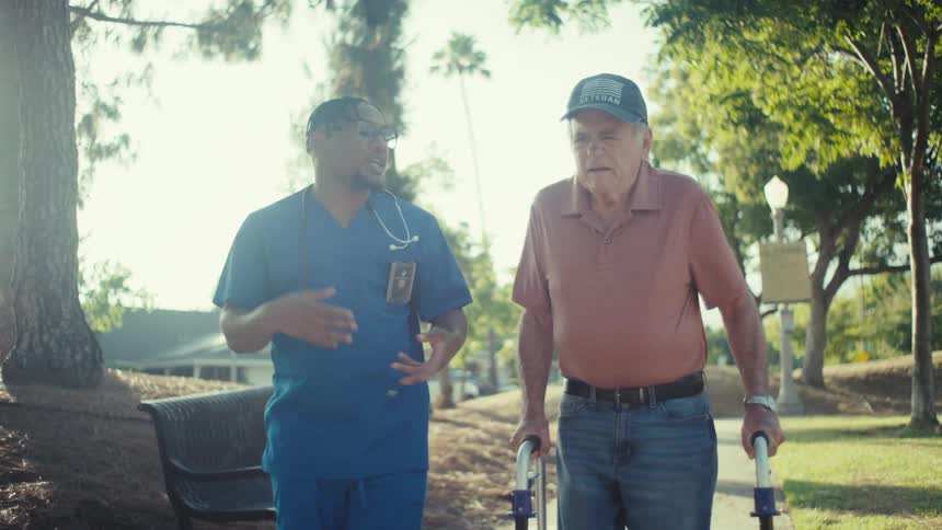 A person in blue scrubs assists an elderly individual using a walker on a paved path in a park with trees and benches