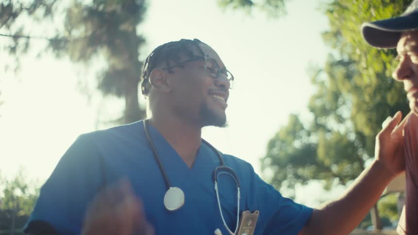 A man in blue medical scrubs and glasses shows him smiling and nodding while holding another mans arm the setting appears to be outdoors with trees visible in the background