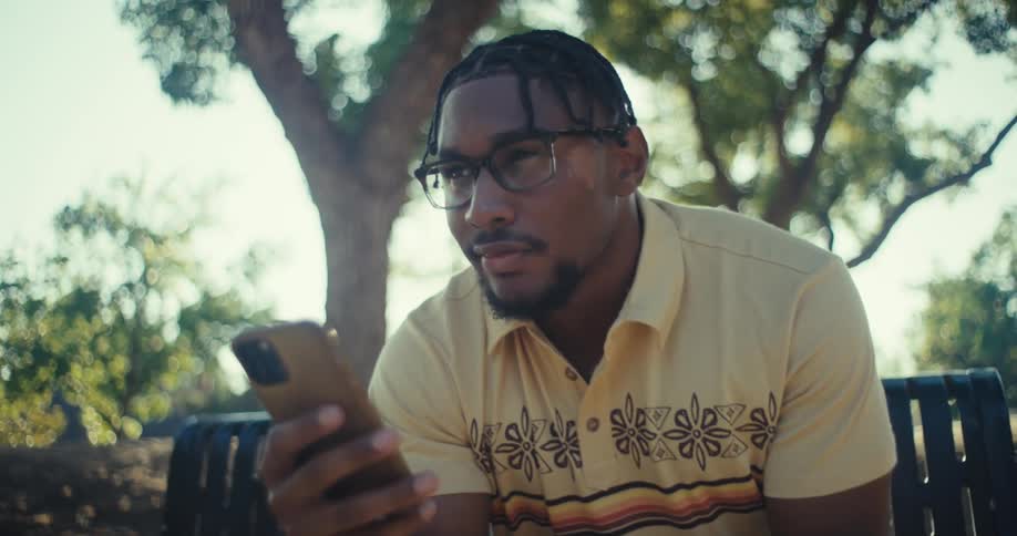 A black man sits on a bench in a sunlit park looking thoughtfully at his phone