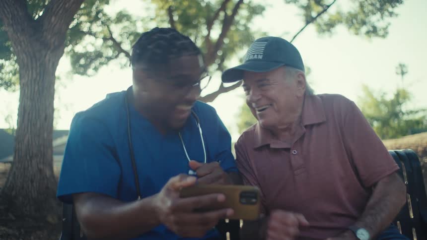Young black doctor and an elderly man react and celebrate happily while viewing media content on a phone as they sit on a bench outdoors during daytime