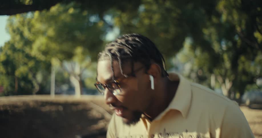 A man with braided hair and glasses wearing a patterned polo shirt smiling and looking at his smartphone outdoors he appears to be walking or enjoying a leisurely moment in a parklike setting