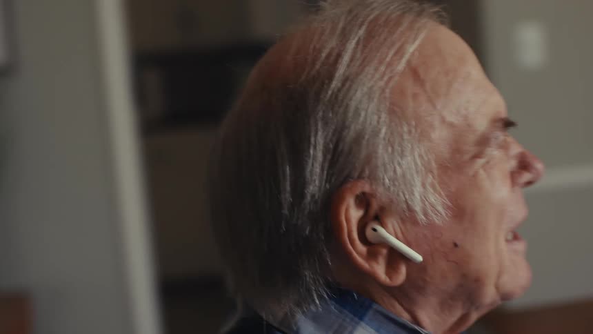 An older person wearing earbuds uses a laptop at a table in a softly lit room with closed curtains engaged in a voice call