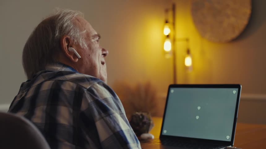 An older man is seated at a table facing away from the camera wearing white wireless earbuds and looking at a laptop screen the room is dimly lit with a decorative lamp providing a warm glow in the background