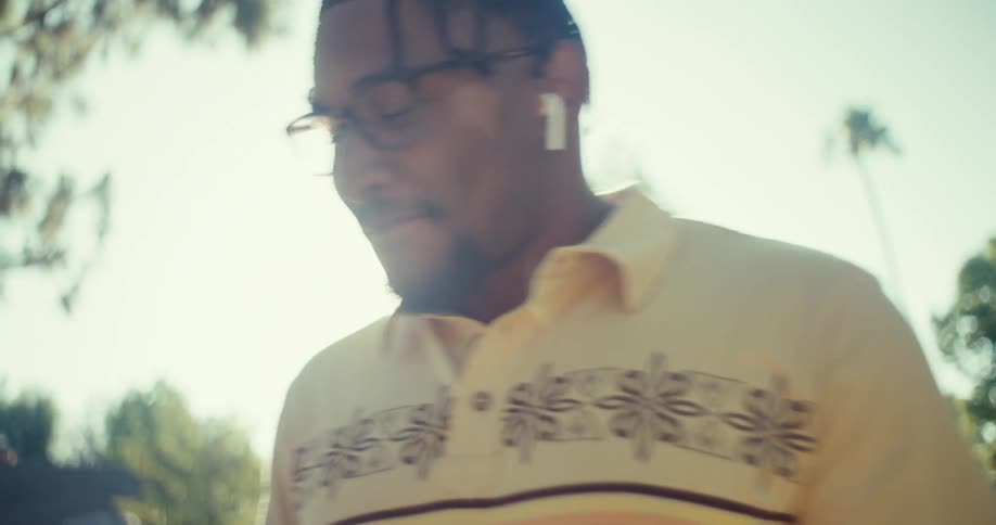 A person wearing a yellow floral shirt walks outdoors speaking on a smartphone while wearing earbuds