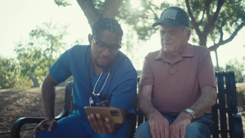 Nurse holding a smartphone and showing it to an elderly man sitting next to him on a bench in an outdoor park