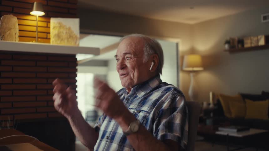 Elderly man makes a phone call with airpods while using a laptop at a table in a living room during daytime