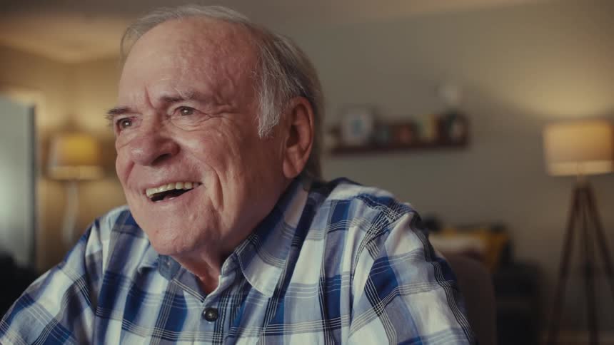 An older man in a plaid shirt sits at a table with a laptop putting on wireless earbuds he then reacts with visible excitement pumping his fists in the air and laughing joyfully