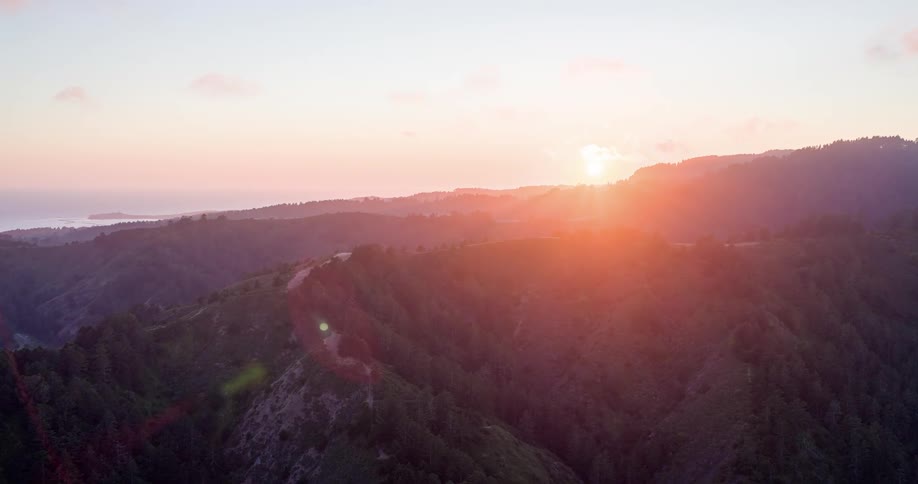 An aerial view captures a vibrant sunset over rolling hills covered in lush green trees with a vast ocean visible on the left horizon