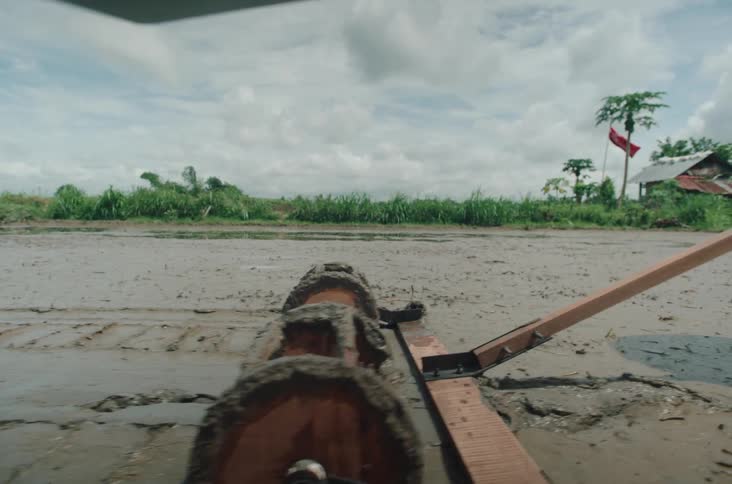A wooden plow dragged by a metal frame carves through a muddy field beneath a cloudy sky with a farmhouse and thick vegetation lining the background