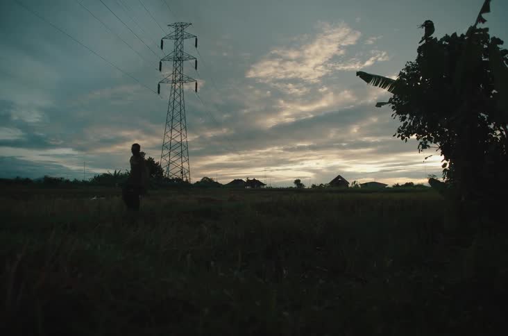 A male walks through a grassy field carrying a bag beneath towering power lines as the sun sets behind distant houses and dramatic clouds