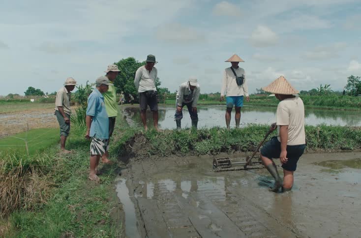 A group of farmers some wearing conical hats gather around a muddy rice field as one man works the soil with a tool
