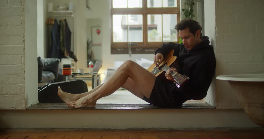 A person sits on the open threshold between bedroom and bathroom playing an acoustic guitar in a cozy room with a couch coat rack and scattered personal items