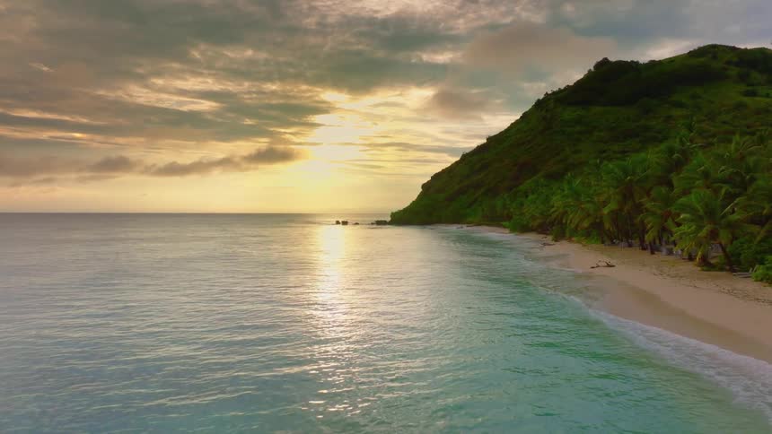 The shoreline stretches with palm trees and a mountain range as light waves hit the shore at sunset