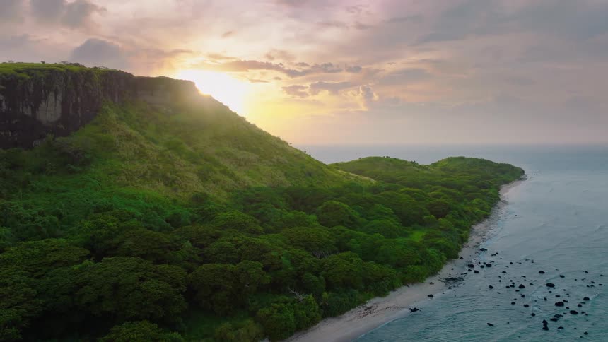 Sunlight filters through hazy clouds over a rugged coastline where jagged cliffs rise above calm shallow waters