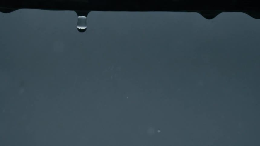 The video shows a dark blurred background with distinct dark horizontal lines possibly representing rooftops or distant trees several raindrops are visible in the foreground falling from an unseen source above the frame