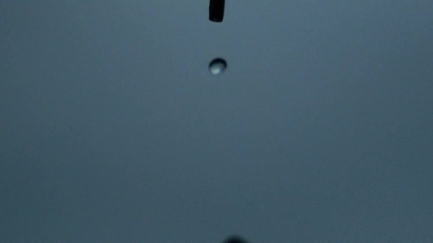 A closeup of a water droplet falling from a small stick against a gray blue stormy blur