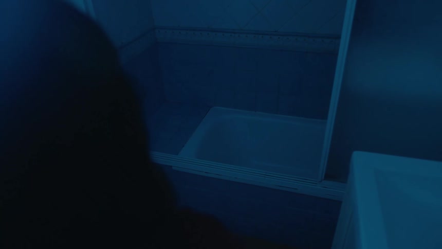 A person stands in a dimly lit blue toned bathroom facing a bathtub